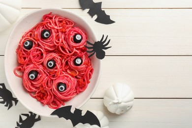 Red pasta with decorative eyes and olives in bowl on white wooden table, flat lay with space for text. Halloween food