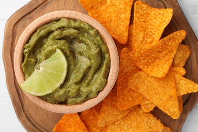 Photo of Bowl of delicious guacamole, lime and nachos chips on white wooden table, top view