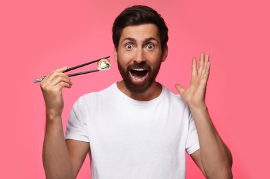 Photo of Emotional man holding tasty sushi roll with chopsticks on pink background