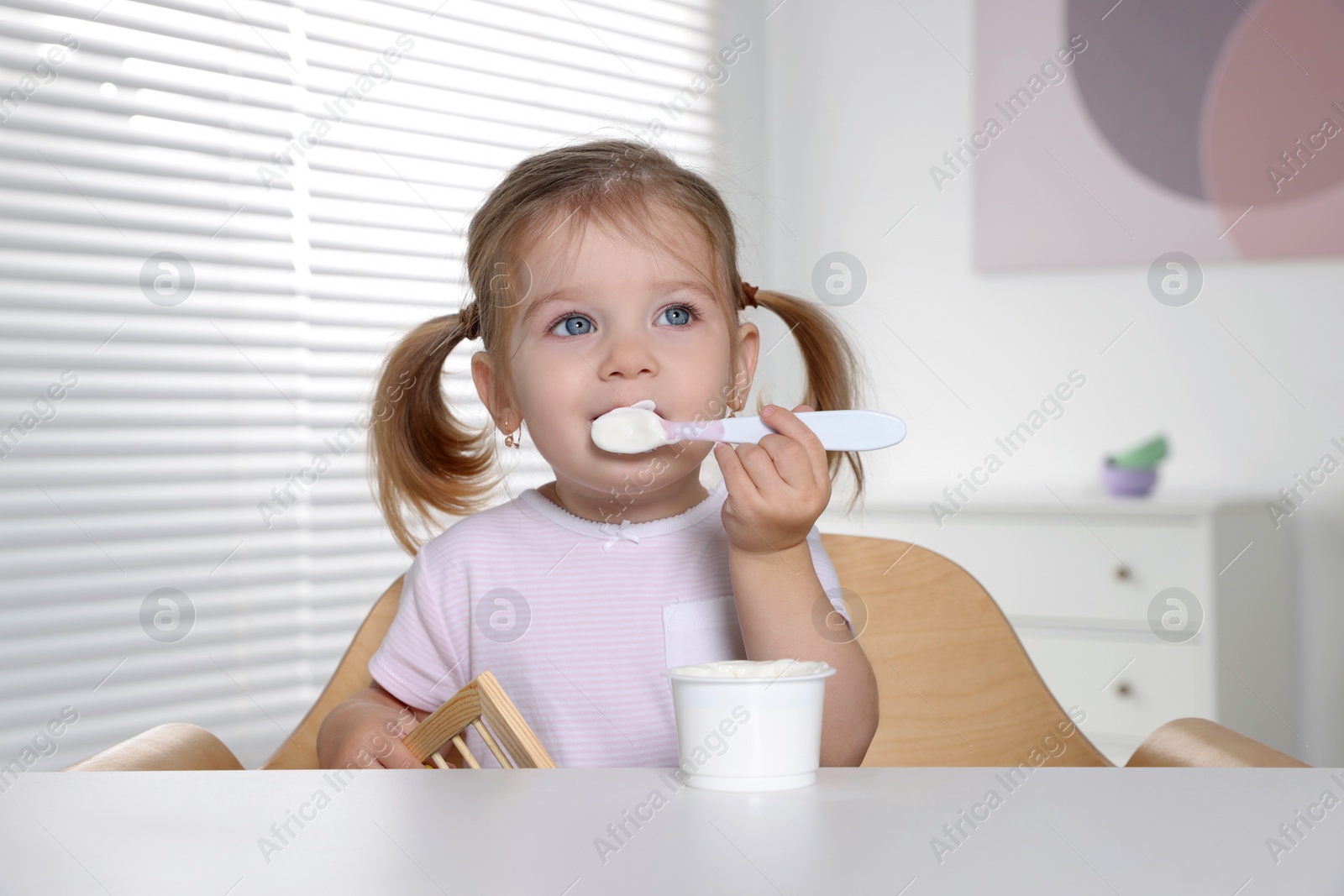 Photo of Cute little child eating tasty yogurt from plastic cup with spoon at white table indoors