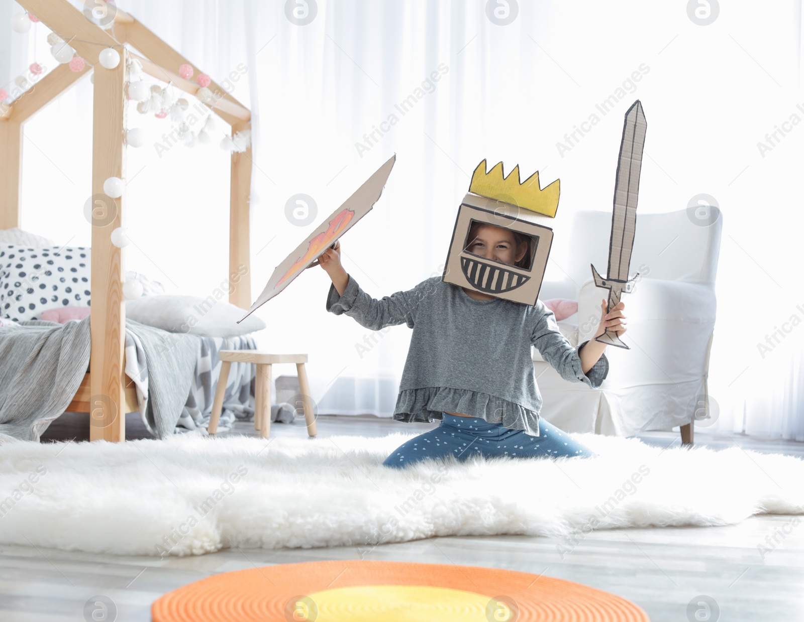 Photo of Cute little girl playing with cardboard armor in bedroom