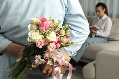 Man hiding bouquet of flowers and present for his beloved woman indoors, closeup