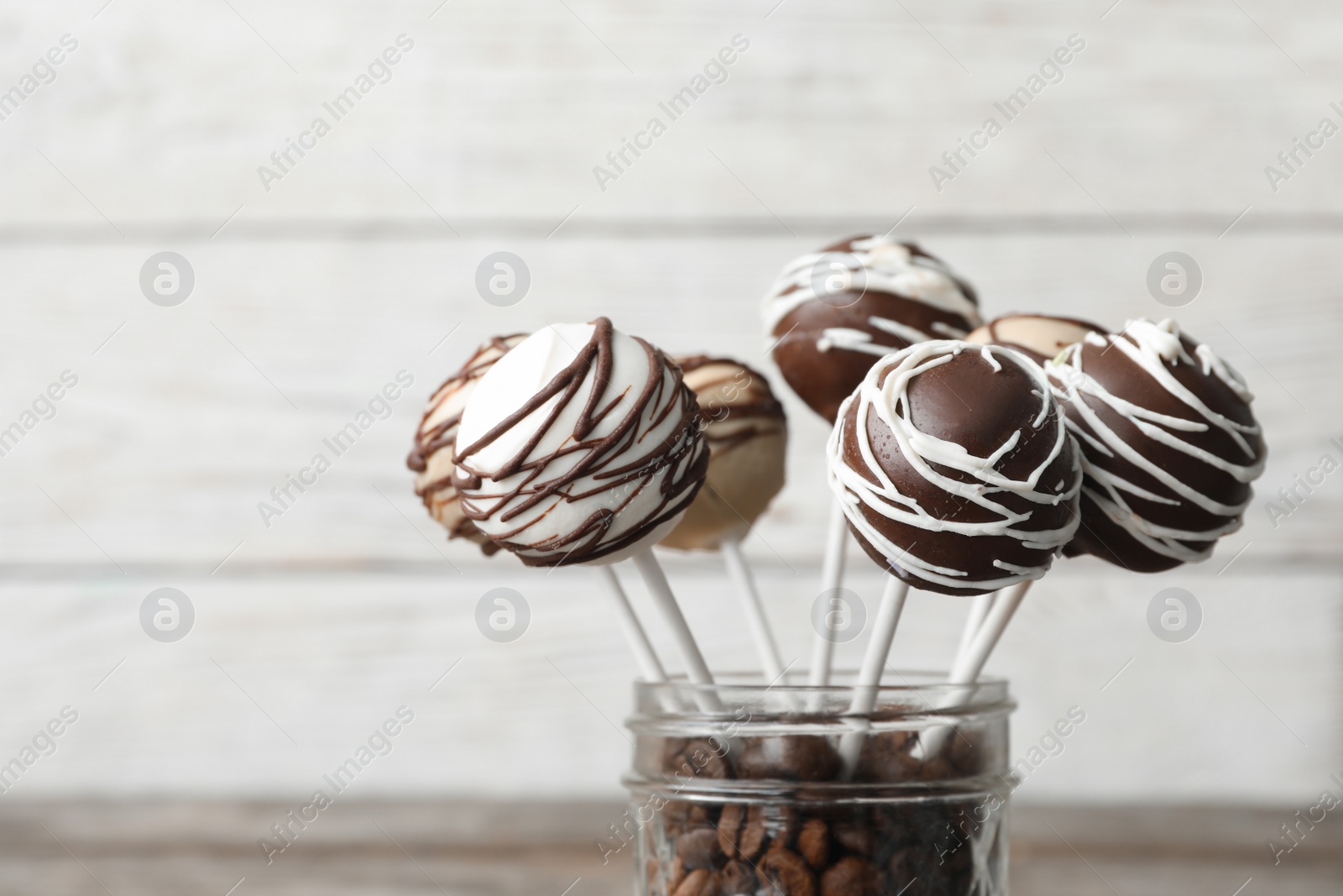 Photo of Yummy cake pops coated with chocolate in glass jar full of coffee beans on blurred background, closeup. Space for text