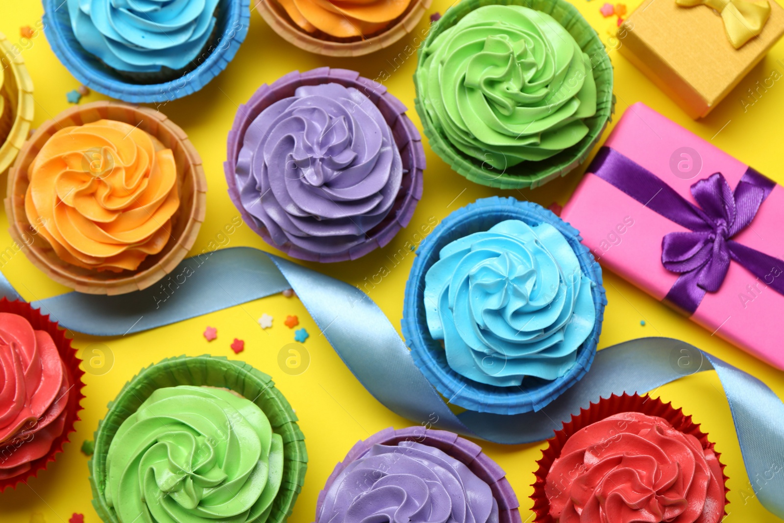 Photo of Delicious birthday cupcakes, gift box, ribbon and sprinkles on yellow background, flat lay