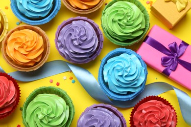 Photo of Delicious birthday cupcakes, gift box, ribbon and sprinkles on yellow background, flat lay