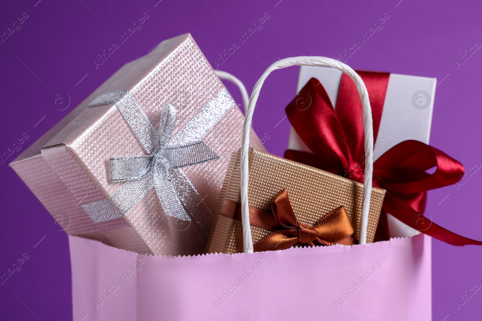 Photo of Pink paper shopping bag full of gift boxes on purple background, closeup