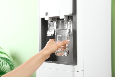 Woman filling glass from water cooler indoors, closeup. Refreshing drink