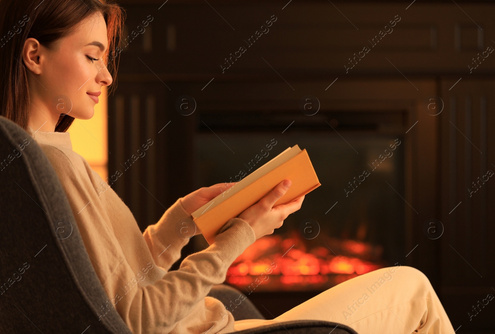 Photo of Young woman reading book near fireplace indoors. Cozy atmosphere