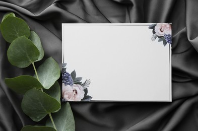 Photo of Blank invitation card and eucalyptus leaves on black fabric, flat lay