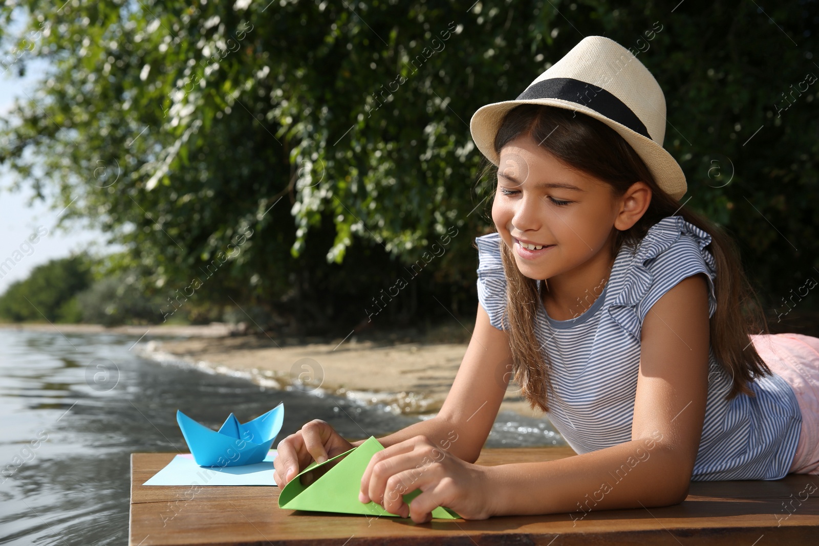 Photo of Cute little girl making paper boats on wooden pier near river