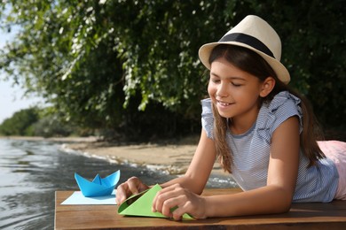 Cute little girl making paper boats on wooden pier near river