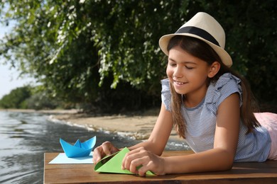 Photo of Cute little girl making paper boats on wooden pier near river