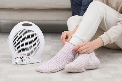 Photo of Woman warming near modern electric fan heater on floor indoors, closeup