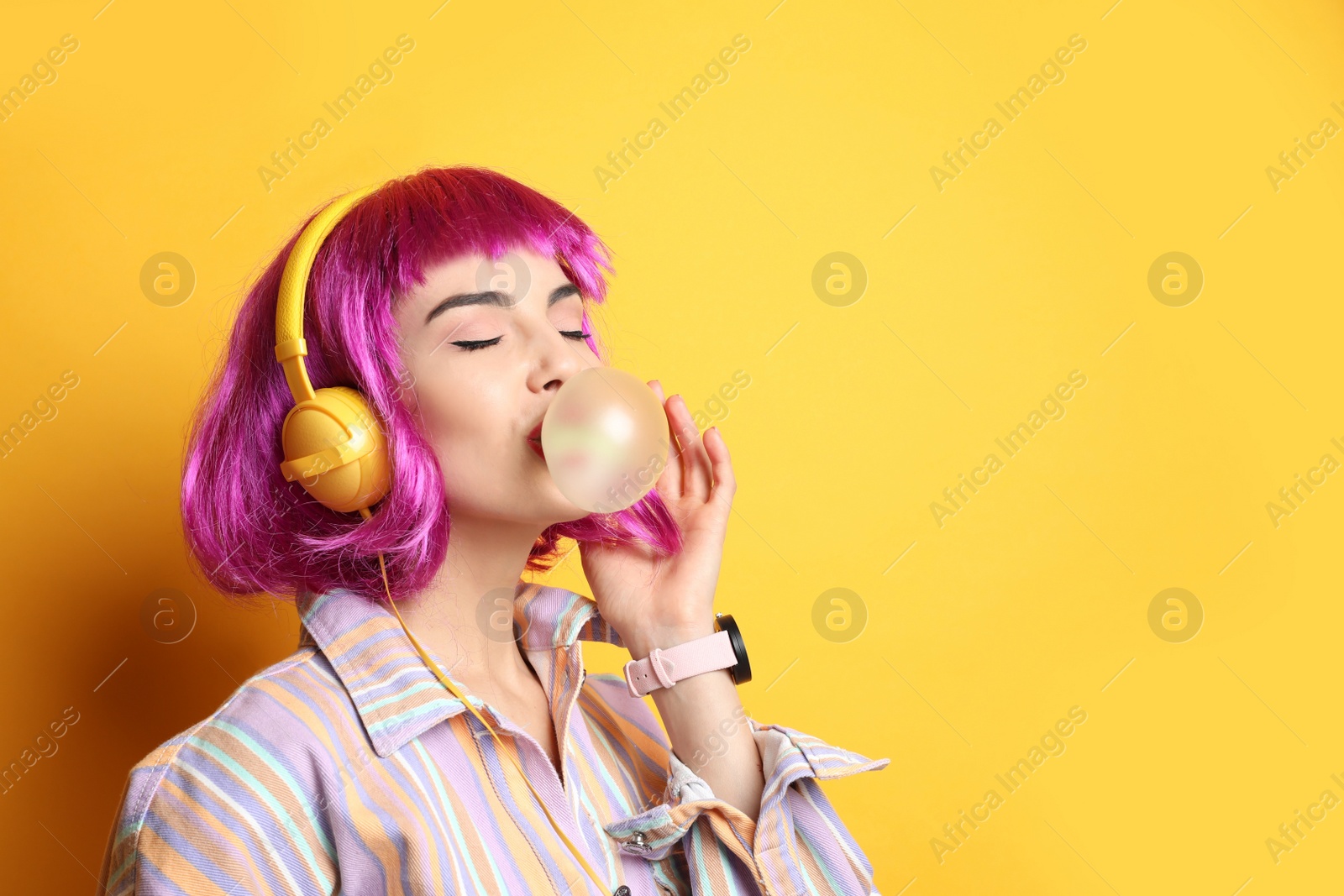 Photo of Fashionable young woman in colorful wig with headphones blowing bubblegum on yellow background, space for text