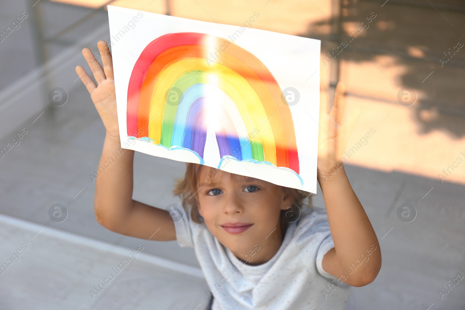 Photo of Little boy with picture of rainbow near window, view from outdoors. Stay at home concept