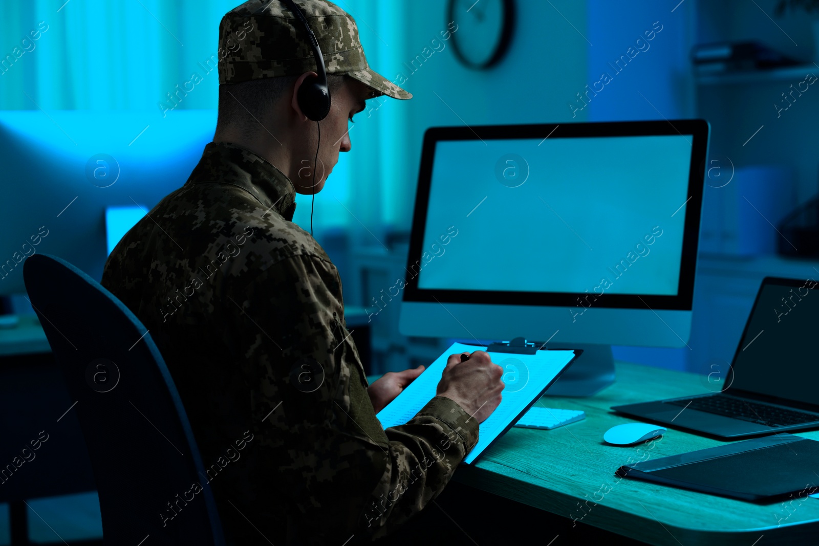 Image of Military service. Soldier with clipboard and headphones working at wooden table in office at night