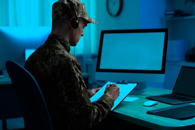 Image of Military service. Soldier with clipboard and headphones working at wooden table in office at night