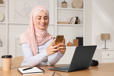Photo of Muslim woman using smartphone near laptop at wooden table in room