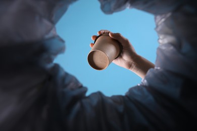 Photo of Bottom view of woman throwing paper cup into trash bin on light blue background, closeup