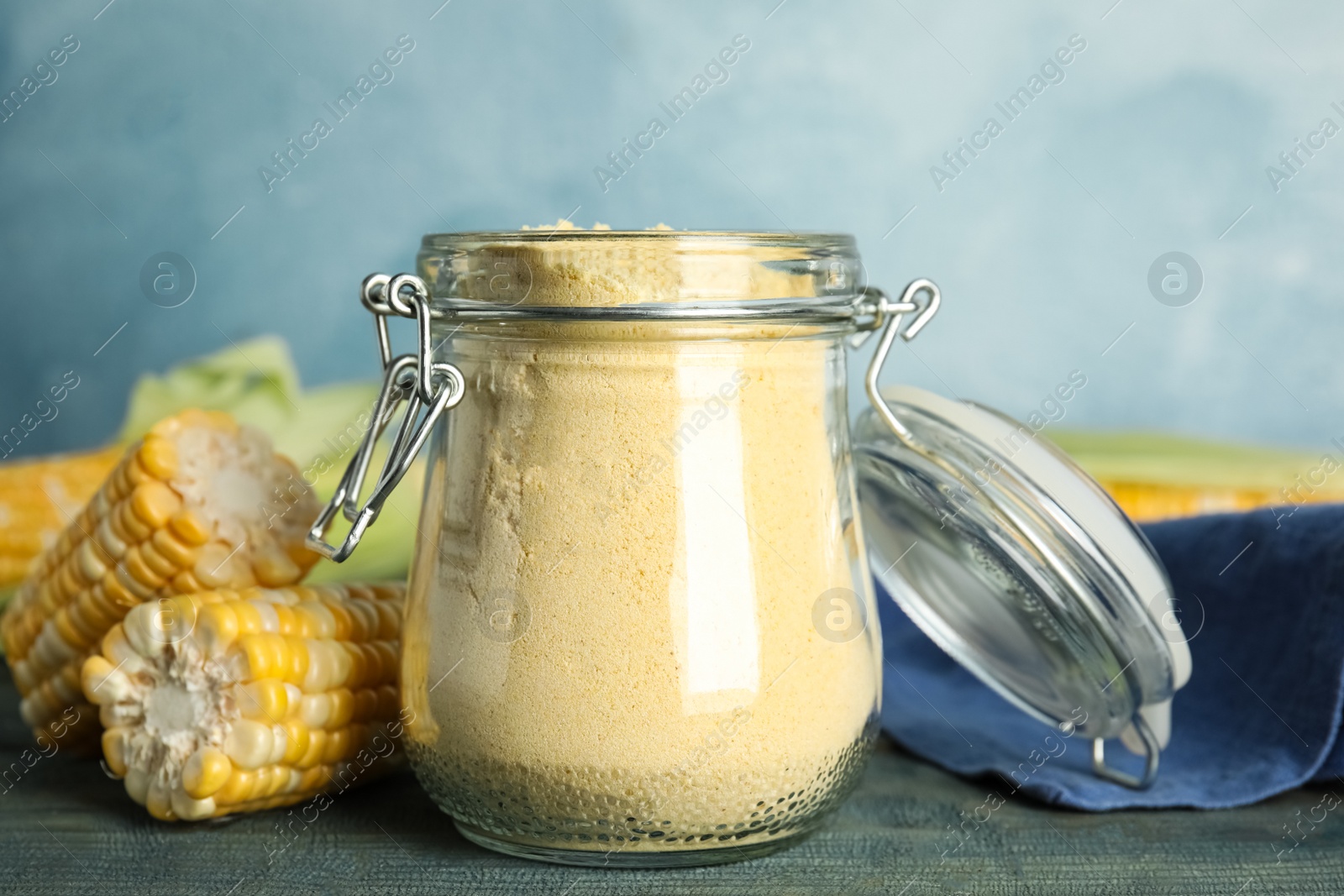 Photo of Corn flour in glass jar and fresh cobs on blue wooden table