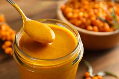 Photo of Spoon with delicious sea buckthorn jam over jar, closeup