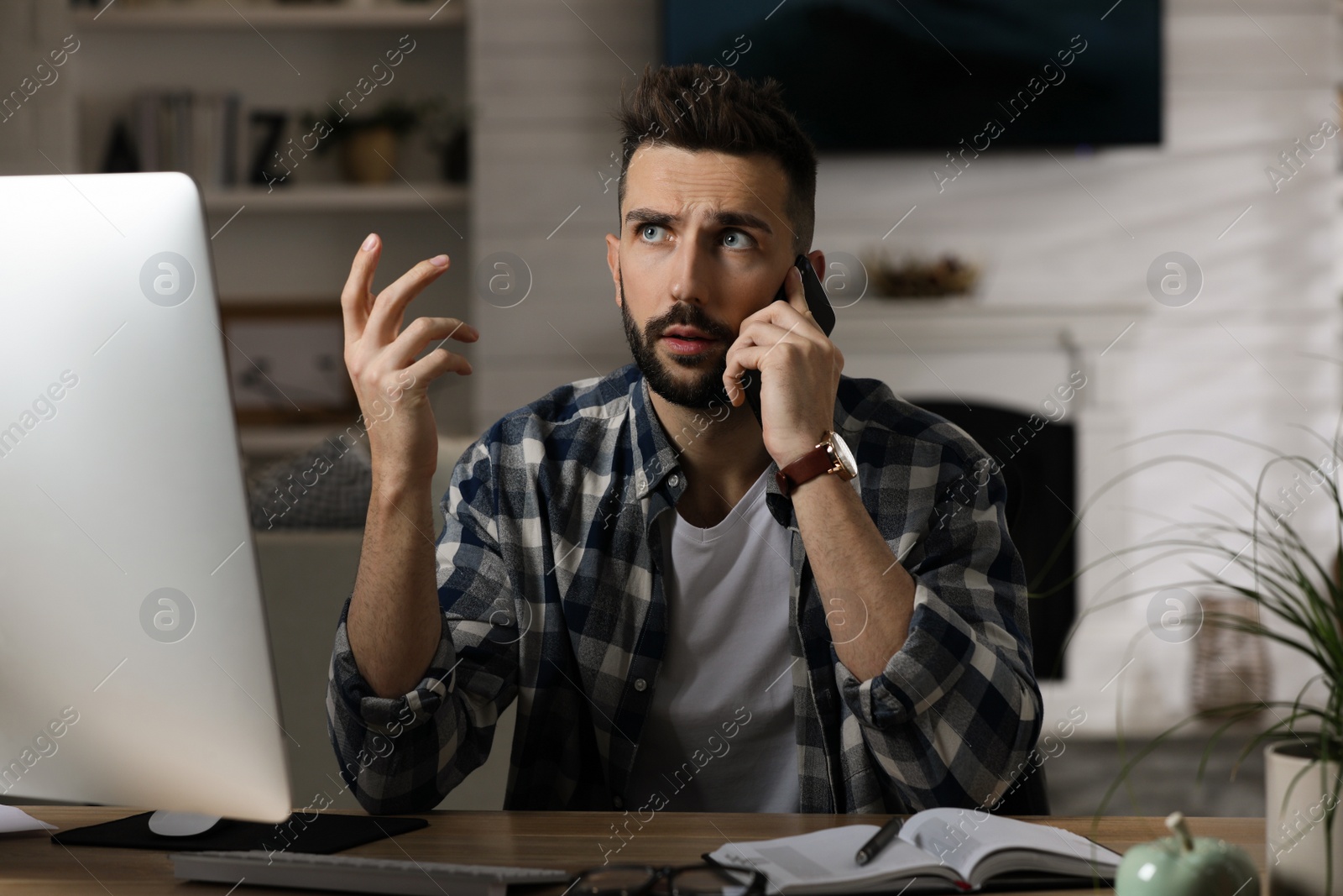 Photo of Man talking on phone while working with computer at table in home office