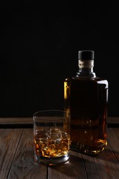 Whiskey with ice cubes in glass and bottle on wooden crate against black background