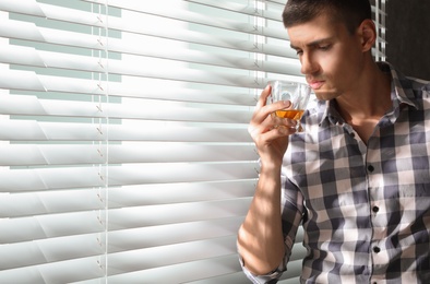 Young man with glass of whiskey near window indoors. Space for text