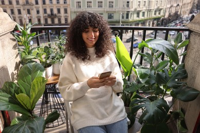 Beautiful young woman using smartphone surrounded by houseplants on balcony