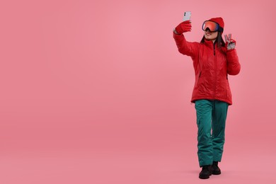 Photo of Happy woman in winter sportswear and goggles taking selfie and showing peace sign on pink background. Space for text