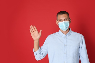 Photo of Man in protective mask showing hello gesture on red background, space for text. Keeping social distance during coronavirus pandemic
