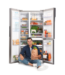 Man with bag of groceries near open refrigerator on white background