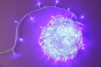 Photo of Glowing Christmas lights on violet  background, top view