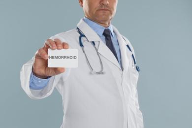 Photo of Doctor holding business card with word HEMORRHOID on light grey background, closeup