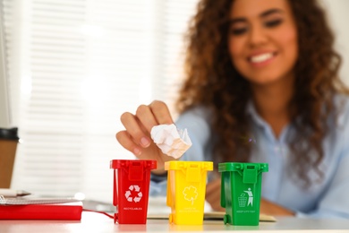 Photo of Young African-American woman throwing paper into mini recycling bin at table in office, focus on hand