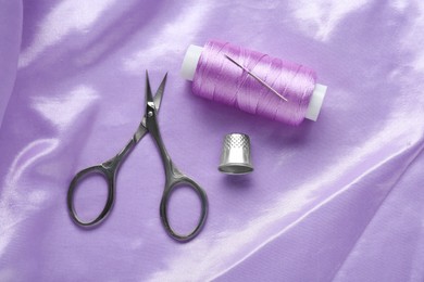 Photo of Thimble, scissors and sewing thread on purple cloth, flat lay. Space for text