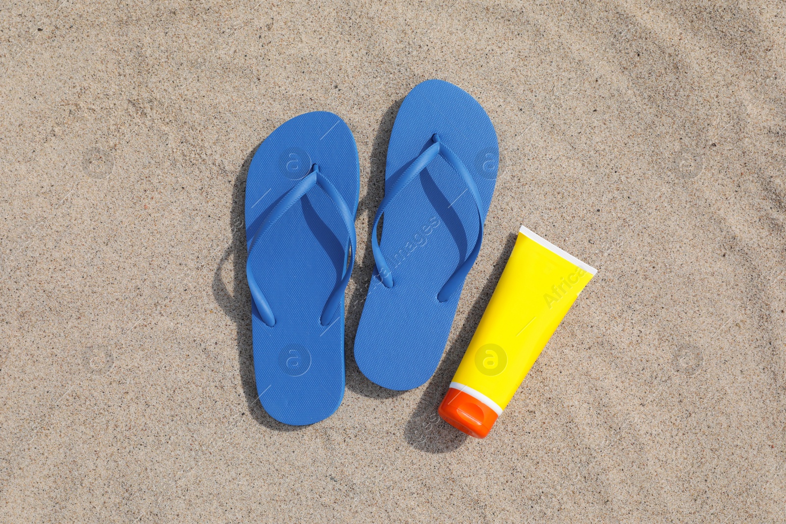 Photo of Blue flip flops and sunscreen on sand, flat lay