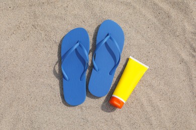 Photo of Blue flip flops and sunscreen on sand, flat lay