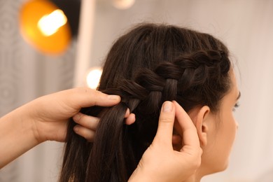 Photo of Professional stylist braiding client's hair on blurred background, closeup