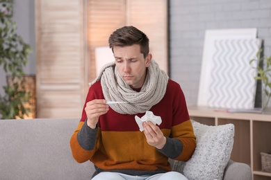 Sick young man with thermometer suffering from cold on sofa at home