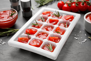 Photo of Ice cube tray with tomatoes, sauce and fresh rosemary on grey table, closeup