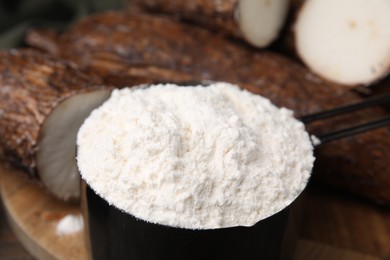 Photo of Scoop with cassava flour and roots on table, closeup