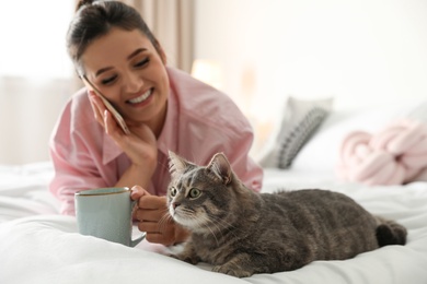 Photo of Young woman with cup of coffee talking on phone while lying near cute cat in bedroom. Pet and owner