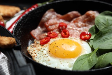 Tasty fried egg with bacon, chili pepper and spinach in pan, closeup