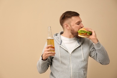 Young man with beer eating tasty burger on color background. Space for text