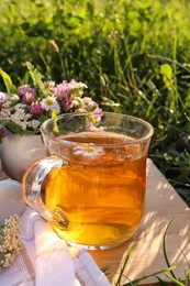 Cup of aromatic herbal tea and ceramic mortar with different wildflowers on wooden board in meadow