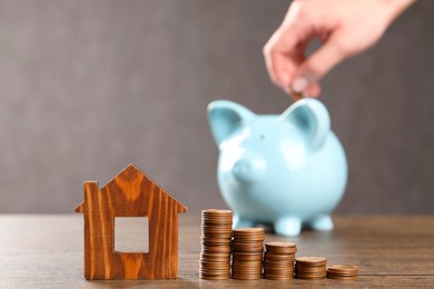 Photo of Woman putting coin into piggy bank at wooden table, focus on house model and money