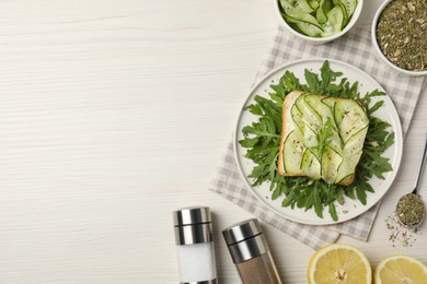 Photo of Tasty cucumber sandwich with seasoning, arugula and lemon slices on white wooden table, flat lay. Space for text