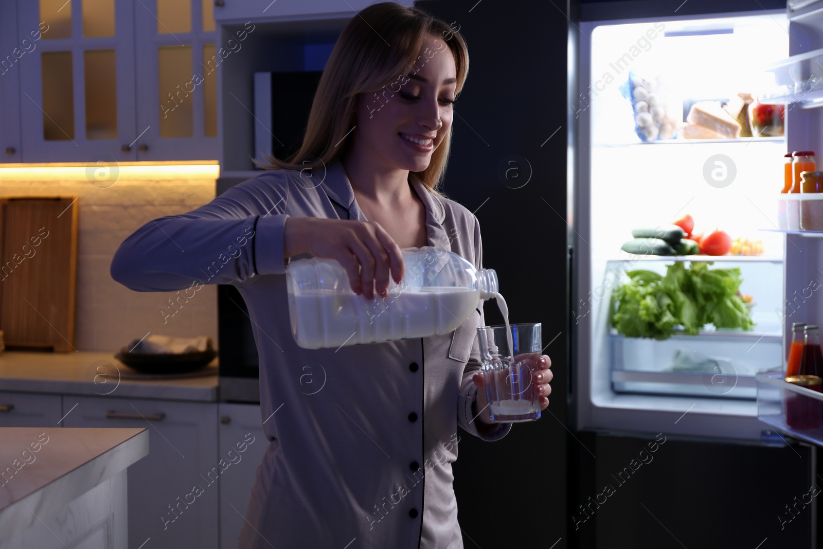 Photo of Young woman pouring milk from gallon bottle into glass near refrigerator in kitchen at night