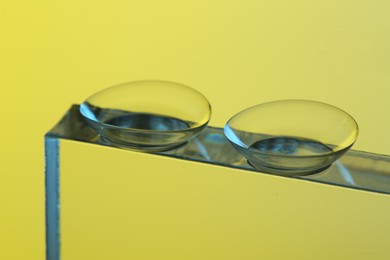 Photo of Pair of contact lenses on glass against pale yellow background, closeup
